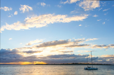 Wall Mural - Catamaran sailboat in water at sunset with sun breaking though clouds on horizon