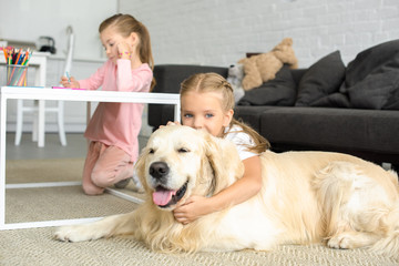 Wall Mural - selective focus of kid hugging golden retriever dog on floor while sister drawing picture at home