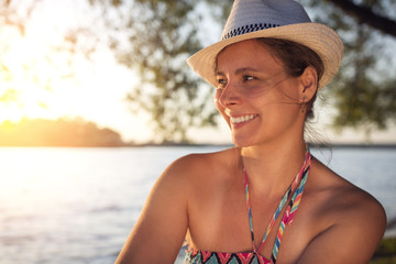 Fototapete - Smiling happy beautiful tanned lady in summer hat on the lake shore against background of sunset over the horizon. Lovely girl on beach.