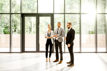 Wall Mural - Group of a young strictly dressed business people standing together with digital tablet at the modern office hall indoors