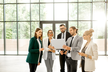 Wall Mural - Group of a young strictly dressed business people standing together with digital tablet at the modern office hall indoors