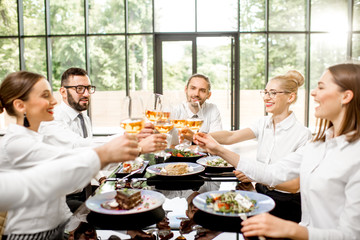 Group of business people dressed in white shirts clinking with wine glasses during a business lunch with delicious meals at the modern restaurant