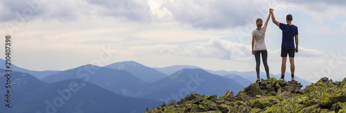 Fotografía Back view of young tourist couple, athletic man and slim girl standing with raised arms holding hands on rocky mountain top enjoying fantastic panorama