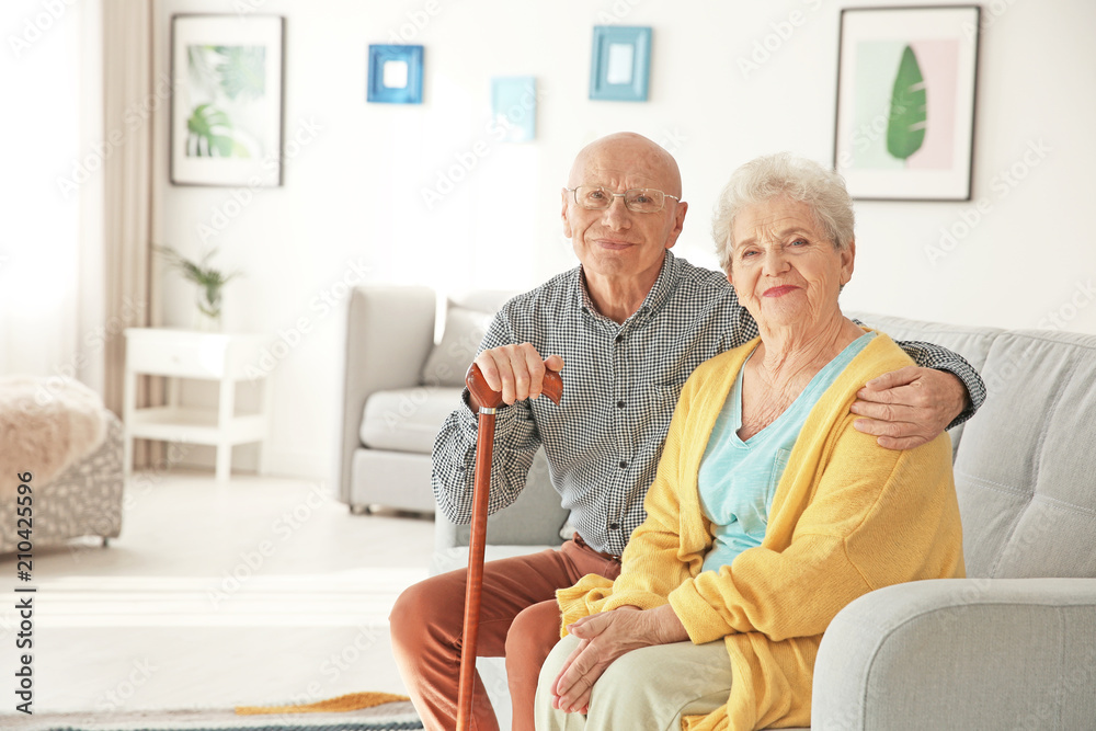 Elderly couple sitting on couch in living room - obrazy, fototapety, plakaty 
