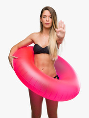 Poster - Beautiful young woman in bikini holding pink float with open hand doing stop sign with serious and confident expression, defense gesture