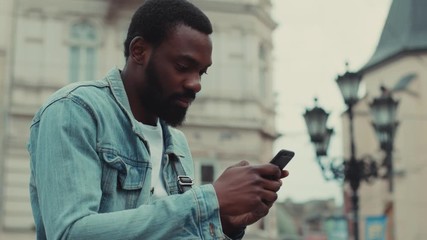 Smiling young African American man uses the phone sitting outdoor at the street business shopping internet face technology fashion call texting mobile smart attractive cell communication slow motion