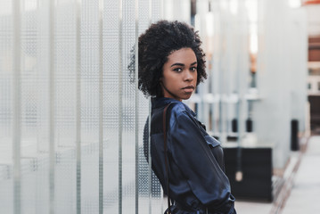 The portrait of beautiful young Brazilian female standing outdoors and leaning against regular glass blocks; charming black girl with curly Afro hair is standing outdoors near the glass installation