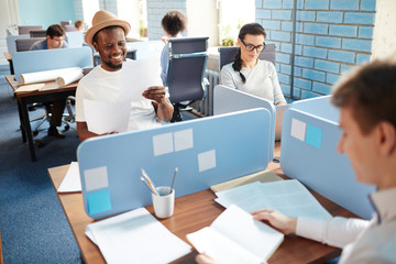 Wall Mural - Happy young African-american businessman in hat and t-shirt sitting by individual workplace in open space office and reading paper among colleagues