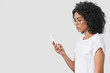 © wayhome.studio  - Beautiful African American female stands in profile, has serious look as reads text message on smart phone, connceted to wifi, surfes internet via modern device, isolated on white concrete wall