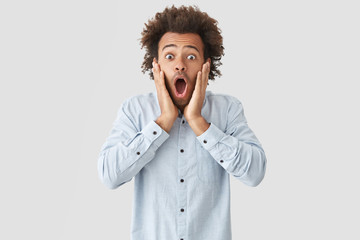 Wall Mural - Horizontal portrait of shocked scared African American guy keeps mouth wide opened and touches cheeks, being terrified as his favourite pet disappeared, isolated over white studio background.