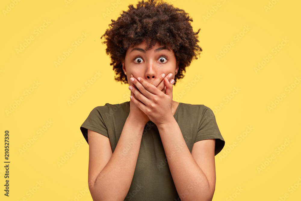 Emotive stupefied young African American female with dark skin, covers mouth as tries to be speechless, looks with popped eyes, stands against yellow background, shocked as notices car accident - obrazy, fototapety, plakaty 
