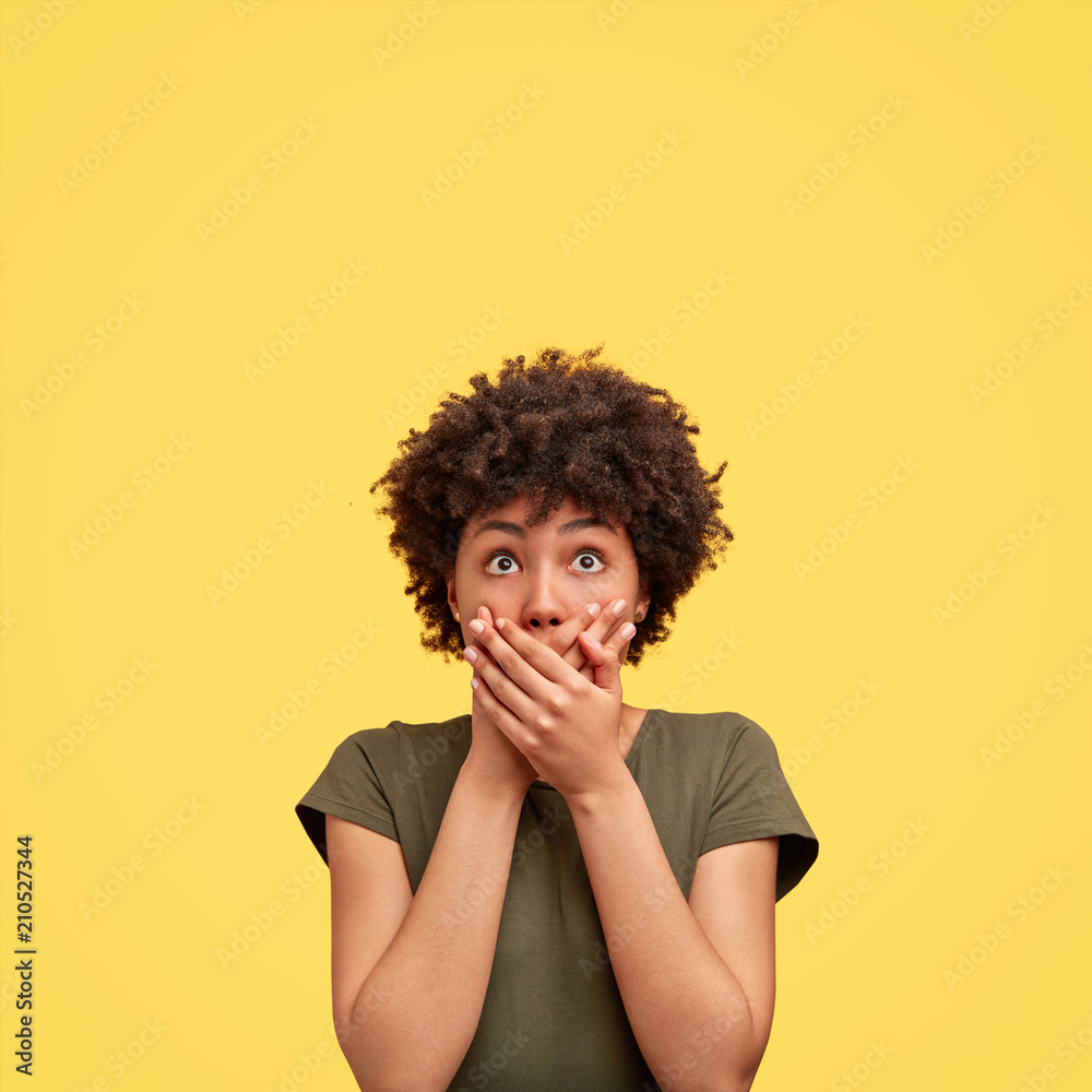 Vertical portrait of emotional scared African American female closes mouth with palms, looks with frightened expression upwards, tries not to make any sound, isolated over yellow background. - obrazy, fototapety, plakaty 