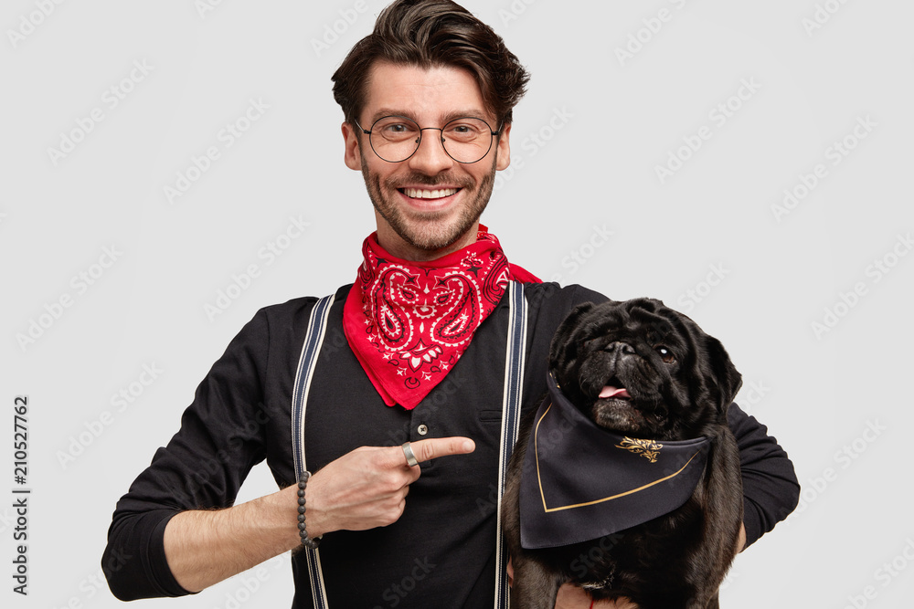 Handsome stylish male vet points at his black pug dog, wears spectacles, has happy expression, poses against white blank wall. Lovely pet in hands of host. Friendship and pedigree animals concept - obrazy, fototapety, plakaty 