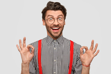 Positive unshaven guy makes ok gesture with both hands, has funny expression, dressed elegantly, approves something, isolated over white background. People, joy, happiness, lifestyle concept