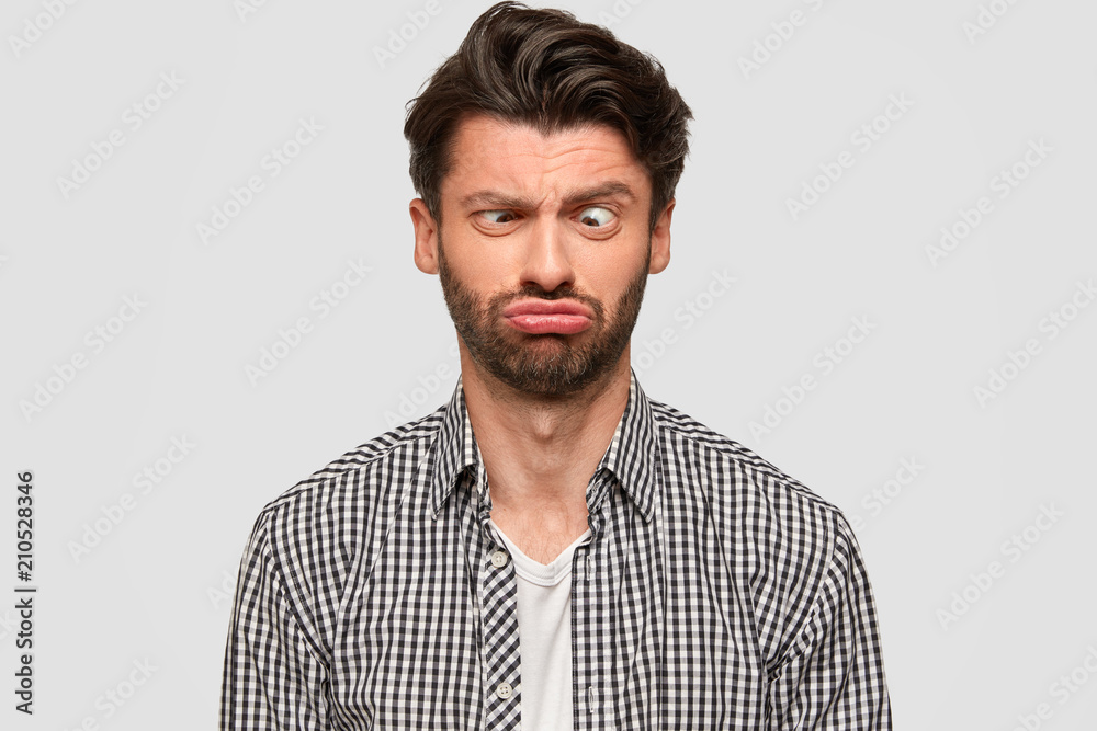 Funny European male makes grimace, purses lips and crosses eyes, foolishes indoor, has fun, dressed in fashionable shirt, isolated over white background. People and facial expressions concept - obrazy, fototapety, plakaty 