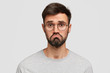 Headshot of attractive young male with beard, purses lips, feels puzzled, has hesitant and miserable look, being disappointed and displeased, dressed casually, isolated over white background.