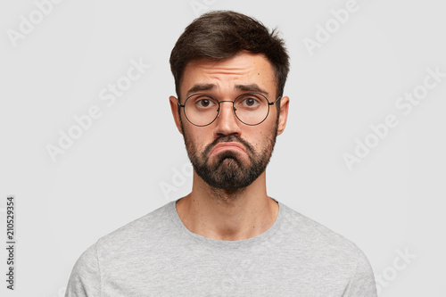 Fotomural Headshot of attractive young male with beard, purses lips, feels puzzled, has hesitant and miserable look, being disappointed and displeased, dressed casually, isolated over white background