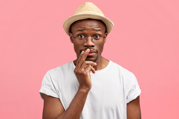 Wall Mural - Puzzed young dark skinned male looks in bewilderment at camera, keeps hand near mouth, wears fashionable hat and casual white t shirt, ready to have walk outside, poses against pink background