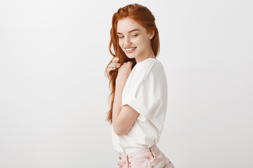 Wall Mural - Studio shot of gentle and feminine woman with natural red hair, standing in profile and gathing with sensual and tender look aside, touching strands of hair and smiling gently, posing over grey wall