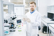 © Seventyfour - Young male microbiologist in laboratory coat standing at desk with microscope and green vegetable samples and holding tablet smiling and looking at camera