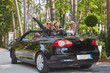 Joyful fashionable female friends raise their hands while sitting in luxury cabriolet car in the park.