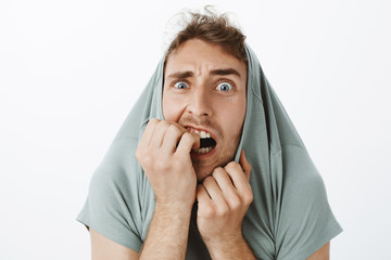 Poster - Indoor shot of funny scared caucasian guy with bristle, pulling t-shirt on head and staring with popped eyes through collar, biting fingernails, trempling from fear and being afraid over gray wall
