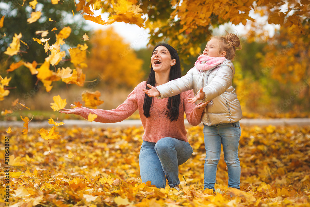 Cheerful girls playing with yellow leaves. Happy mother and little child in the fall time having fun - obrazy, fototapety, plakaty 