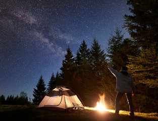 Wall Mural - Camping night in mountains. Brightly lit by campfire tourist tent on pine trees background and silhouette of male hiker pointing at Milky Way in beautiful clear bright blue starry sky. Tourism concept