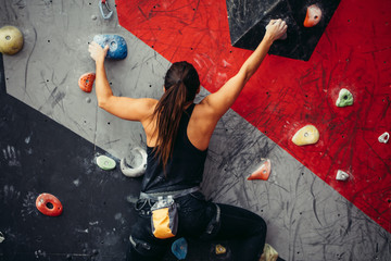 Attractive sporty female climber hanging on handles, studs and hooks of colourful artificial boulder wall in fitness gym, rear view.