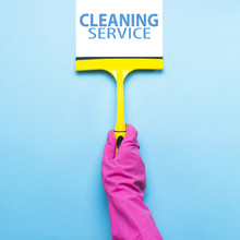 Female Hand In Pink Gloves Holding A Scraper For Cleaning Windows And Removes Dirt From The Surface On A Blue Background. Added Text Cleaning Service. The Concept Of Cleaning The Service