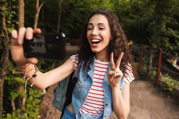 Sticker - Photo of summer teen woman 18-20 wearing casual clothes, showing peace sign and taking selfie photo on mobile phone while walking in green park