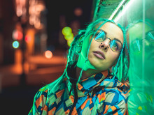 Young Pretty Girl With Unusual Hairstyle Near Glowing Green Neon Light Of The City At Night. Dyed Blue Hair In Braids. Pensive Hipster Teenager In Glasses.