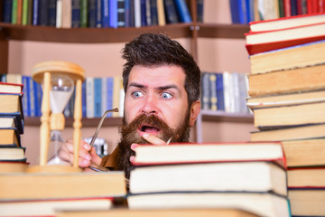 Leinwandbilder - Man, scientist looks at hourglass. Teacher or student with beard studying in library. Man on serious and shocked face watching time is going over, bookshelves on background. Time flow concept.