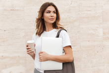 Photo Of Pretty European Business Woman Walking Against Beige Wall Outdoor With Silver Laptop, And Takeaway Coffee In Hands