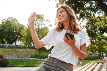 Poster - Photo of affable friendly woman sitting on bench in green park on summer day, and waving hand aside saying hello while holding mobile phone