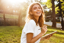 Photo Of Joyful Woman 20s With Broad Smile Sitting On Green Grass In Park With Legs Crossed During Summer Day, And Writing Down Notes With Pen