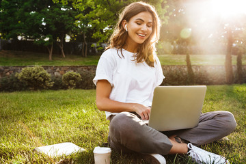 Portrait of pretty young woman sitting on green grass in park with legs crossed during summer day, while using laptop and wireless earphone for video call