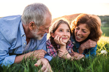 Senior Couple With Granddaughter Outside In Spring Nature, Relaxing On The Grass.