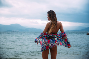 Wall Mural - Rear view of a sexy slender model with long hair stands on the shores of the azure sea in a swimsuit and beach shirt, flattened from the shoulder. The fabric of the blouse neatly covers the girl's ass