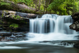 Fototapeta Krajobraz - A forest waterfall with motion, large stones and green trees behind.