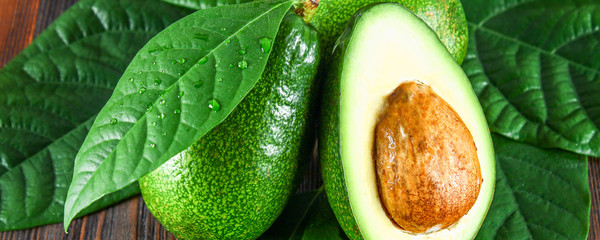 Three green raw ripe avocado fruits and a cut half with a bone with leaves on a brown wooden table.
