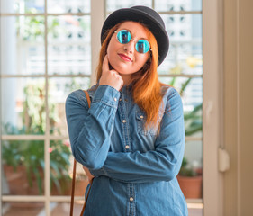 Sticker - Stylish redhead woman wearing bowler hat and sunglasses with hand on chin thinking about question, pensive expression. Smiling with thoughtful face. Doubt concept.