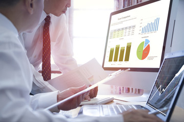 Two Asian Businessmen analysing on business loan comparison on desktop and notebook computer with colorful pie and bar graphs.