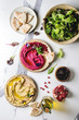Variety of homemade traditional and beetroot spread hummus with pine nuts, olive oil, pomegranate served on ceramic plates with pita bread and green salad on white marble background. Flat lay, space.