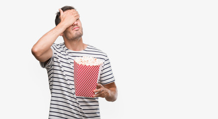 Canvas Print - Handsome young man eating popcorn stressed with hand on head, shocked with shame and surprise face, angry and frustrated. Fear and upset for mistake.