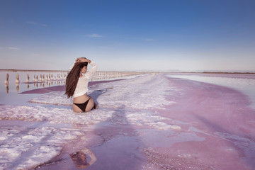 Wall Mural - Beautiful free sexy girl in white swimwear sitting on salty beach at sunset. Sensual tanned woman with long healthy hair posing on salt pink lake enjoying nature landscape. Luxury summer vacation.