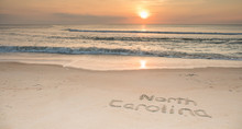 The Words North Carolina Etched In The Sand At Sunrise With Waves Coming Ashore.