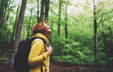 Fototapeta Na ścianę - smiling tourist traveler with backpack into road at summer green forest, girl hiker in yellow hoody looking and enjoying the breath of fresh air in trip, relax holiday concept, blurred background