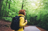 Fototapeta Na ścianę - tourist traveler with backpack into road at summer green forest, girl hiker in yellow hoody looking and enjoying the breath of fresh clean air in trip, relax holiday concept, blurred background