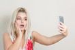 Surprised blonde woman making selfie on mobile phone against a grey background
